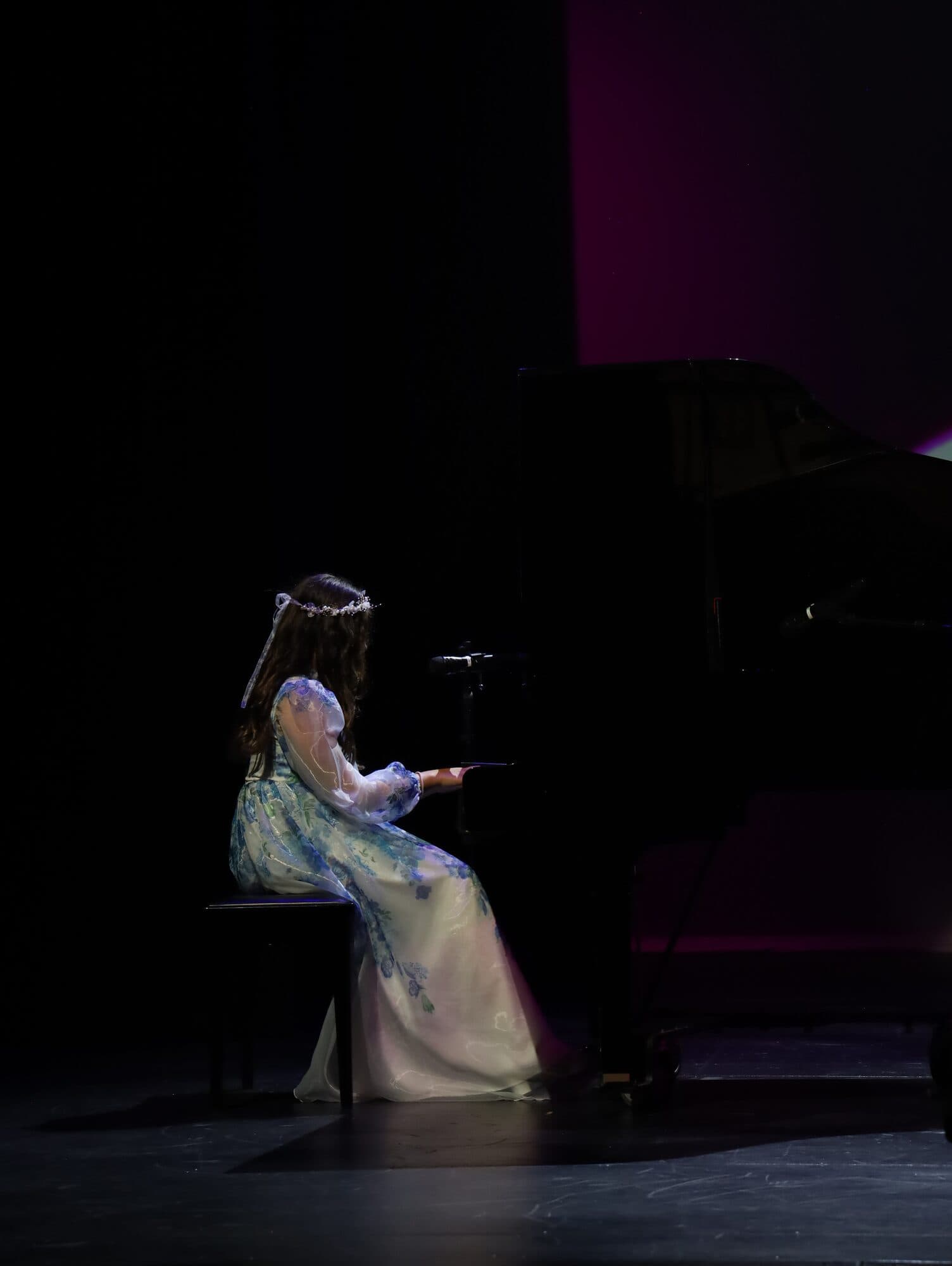 A girl in a floral gown plays a grand piano alone on stage