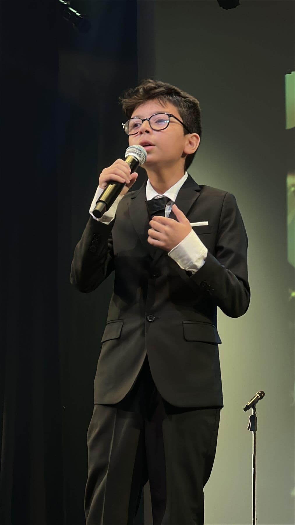 A boy in a black suit sings, his whole body in the frame