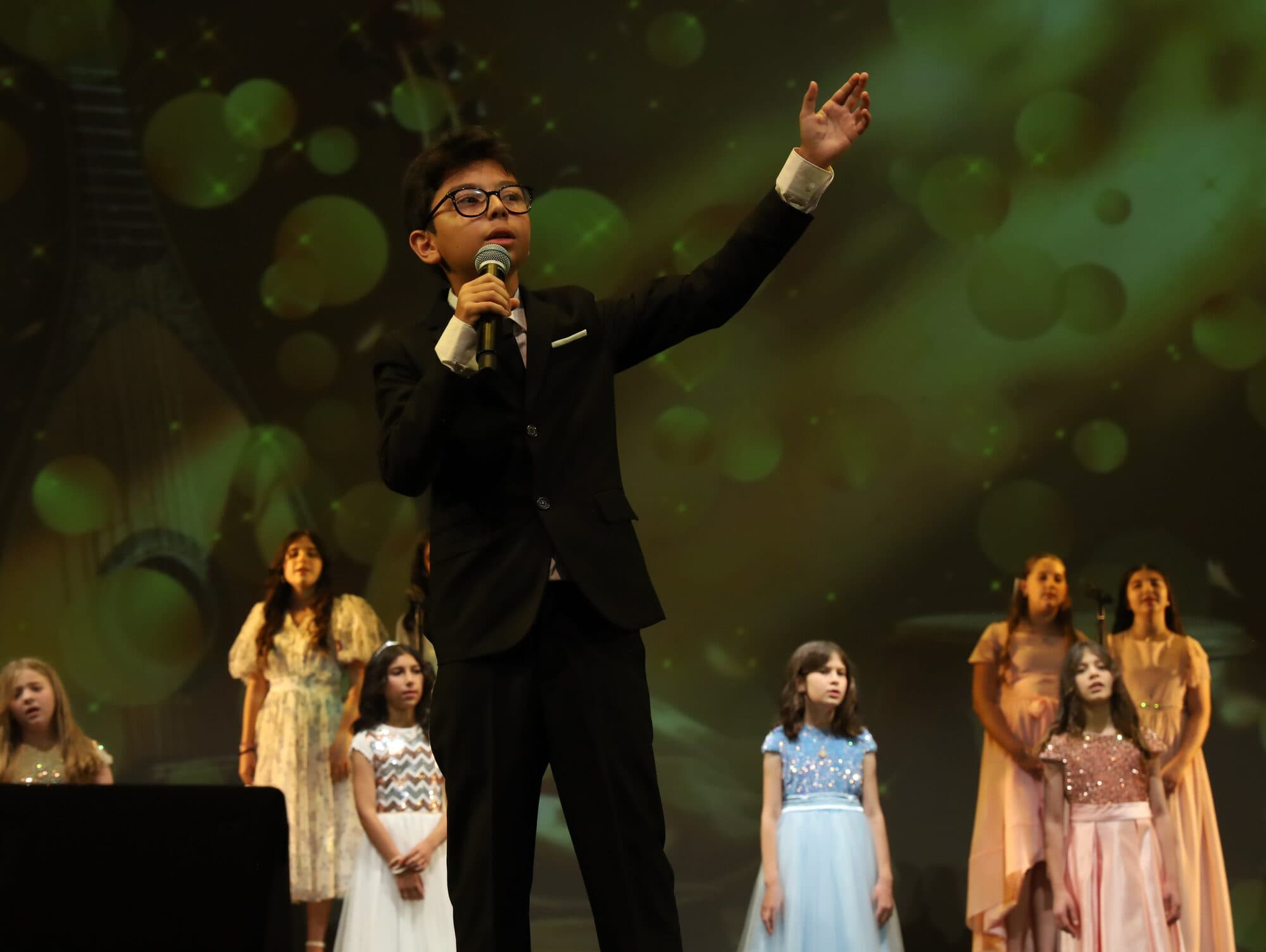 A young soloist in a suit raises his arm as a choir stands behind him